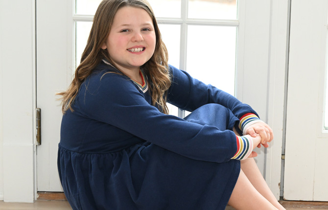 A young girl sits, looking at the camera and smiling.