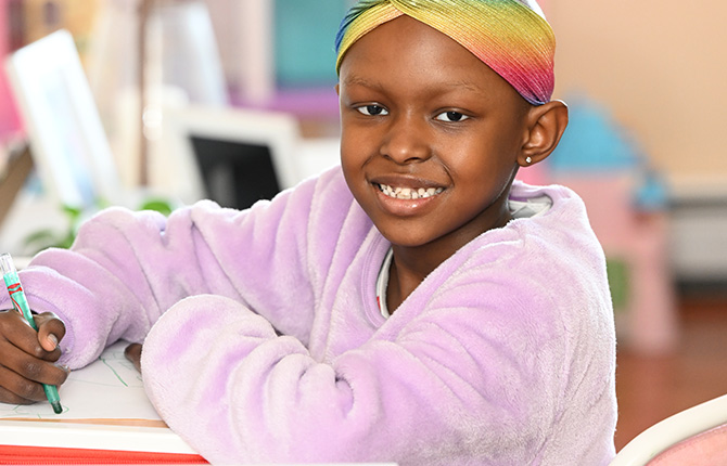 A young girl, coloring, looks at the camera, smiling.