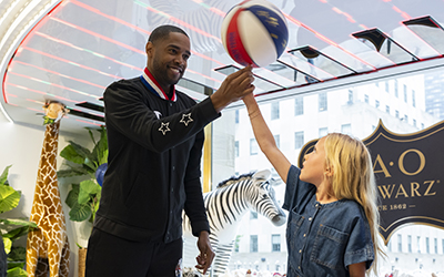 A member of the Harlem Globetrotters showing a young attendee of the NYC for MSK kickoff breakfast at FAO Schwarz how to spin a basketball on her fingertip. 