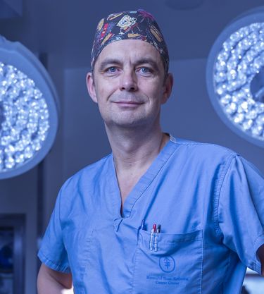 Surgeon in blue scrubs and cap, standing under bright surgical lights.